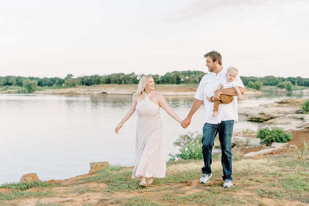 Parents holding hands walking by lake while father holds toddler in Murrell Park summer family session