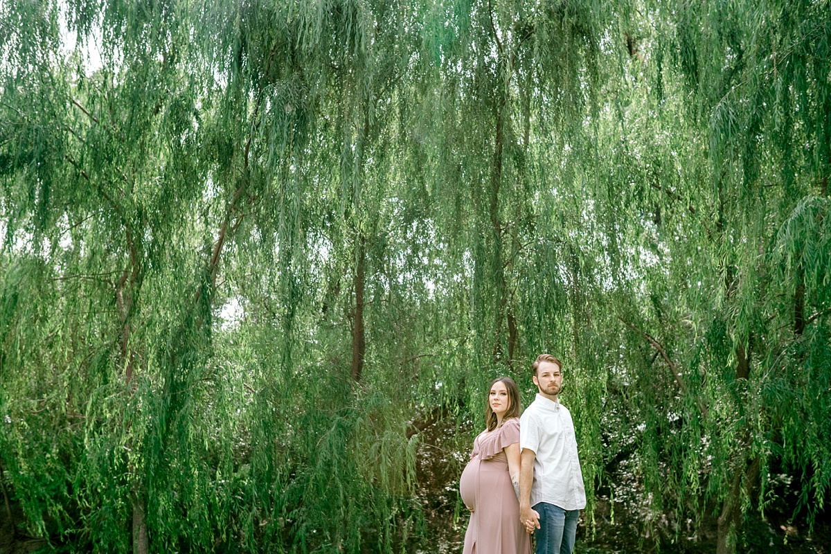 couple holding hands and standing back to back under willow tree Texas maternity session