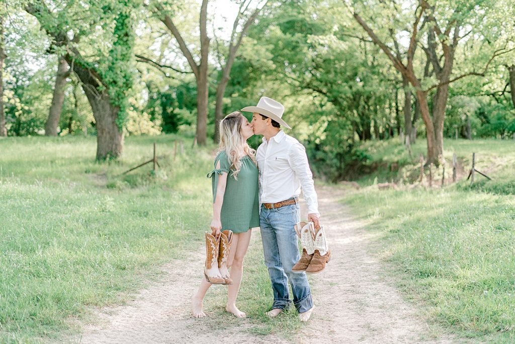 Couple kiss in middle of tree lined dirt road barefoot holding boots