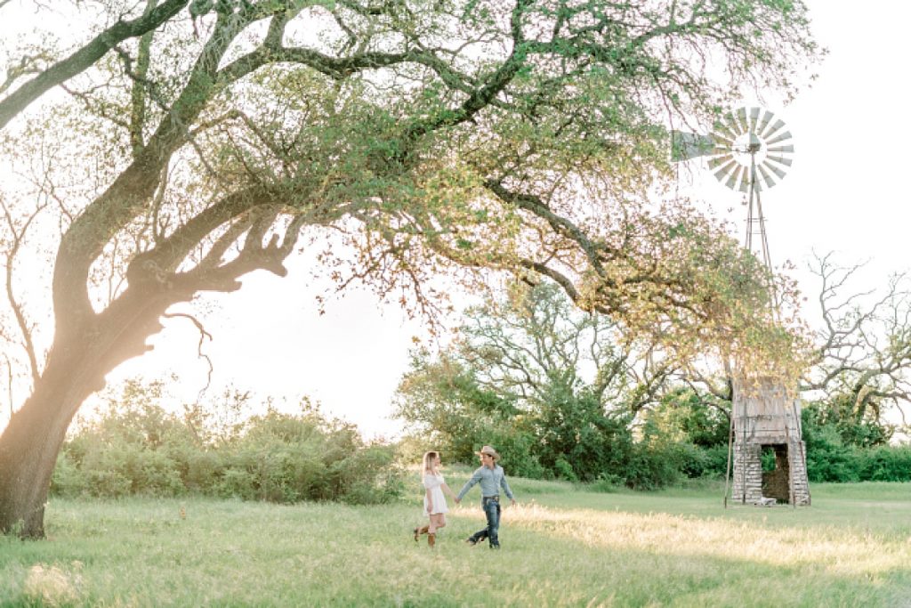 Guy and girl walking under giant tree and rustic windmill| | Hamilton/Hico Engagement 