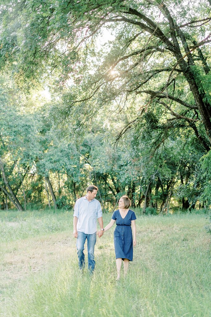 Couple walks hand in hand through meadow with large tree overhead at sunset during their Airfield Falls Engagement Session 