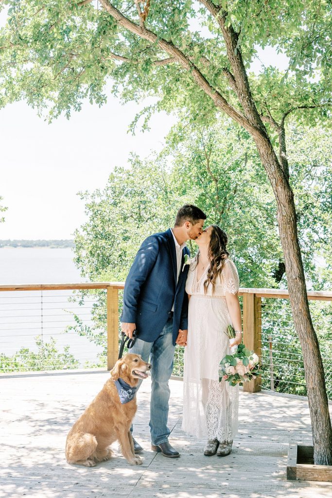 Bride and groom kissing under tree holding dog on leash