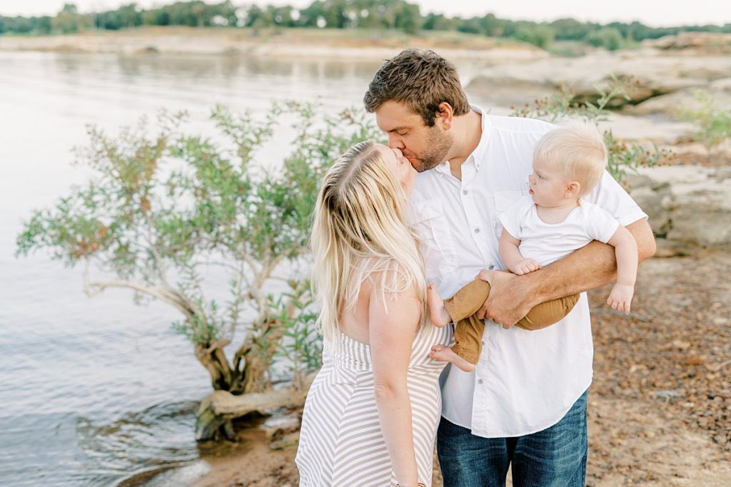 Father holding toddler and kissing wife by lake in Murrell Park summer family session